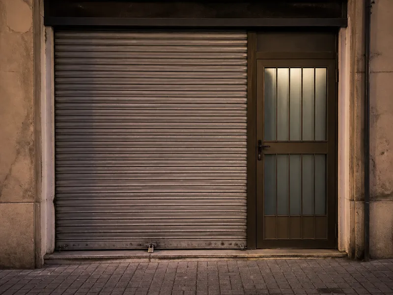 Metal shutter and pedestrian shop door, empty pavement, no lettering.